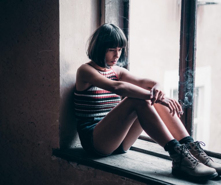 woman sitting by a window smoking looking depressed